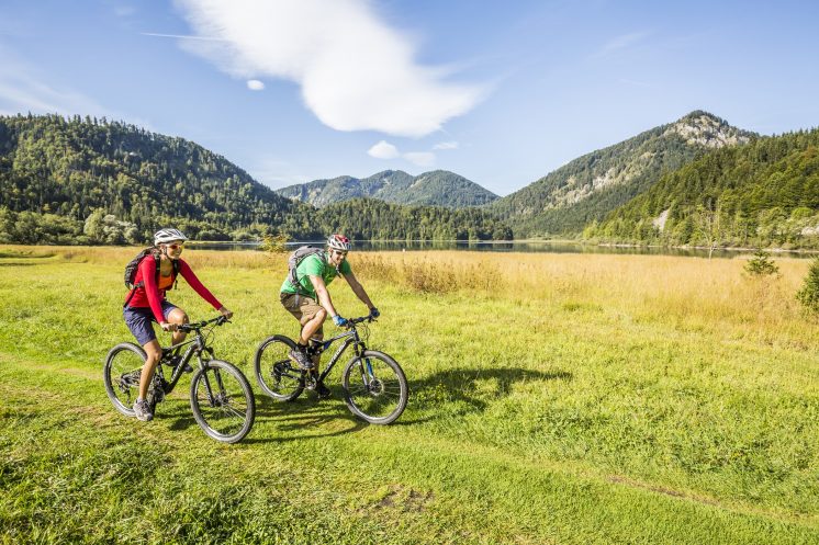 Zwei Fahrradfahrer fahren während ihres Aktivrulaubs in Bayern durch eine grüne Wiese, im Hintergrund die Berge