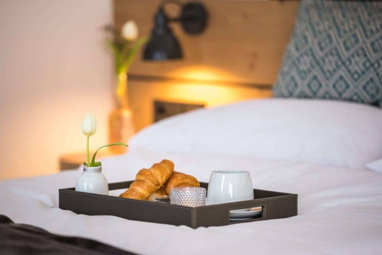 helds-vitalhotel-ruhpolding-fruehstueck-im_zimmer Frühstück mit Croissants, Kerze und Kaffee auf einem Tablett serviert auf einem gemütlichen Hotelbett.