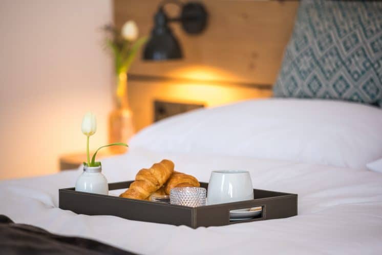 Schön angerichtetes Tablett mit Croissants auf einem weißen Bett, im Hintergrund Blumen auf dem Nachttisch.
